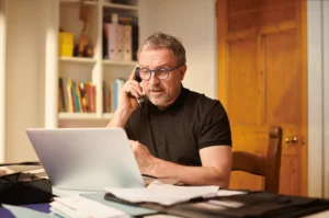 Un homme avec une barbe est au téléphone pour régler des soucis, devant son ordinateur, et son bureau plein d'administratif